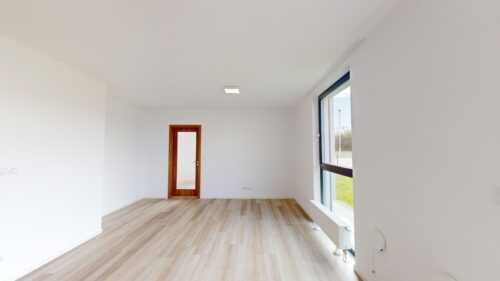 Bright, empty room with white walls, light wood floor, a wooden door with frosted glass, and a large window letting in sunlight.