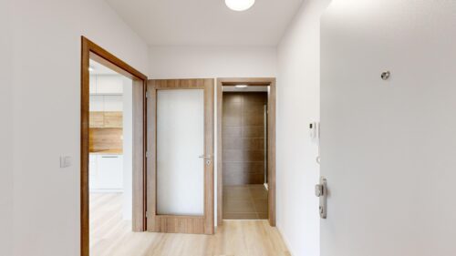 Interior hallway with light wood flooring and white walls; multiple wooden doorways, one open to a tiled bathroom on the right and a kitchen visible to the left.