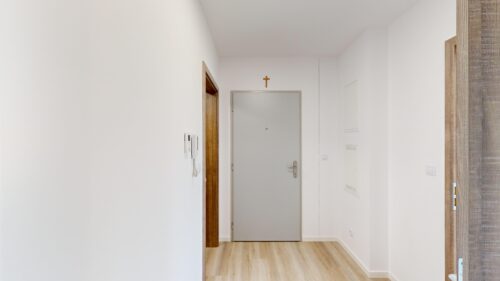 White hallway with light wood flooring, doors on both sides, and a small cross mounted above the door at the end of the hall.