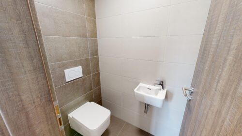 Small modern bathroom with a wall-mounted white toilet, a compact white sink, and gray-brown tiled walls; wooden door frame visible to the left.