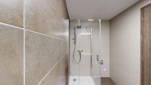 Modern bathroom with glass shower enclosure, chrome fixtures, beige wall tiles, and a wooden door on the right.