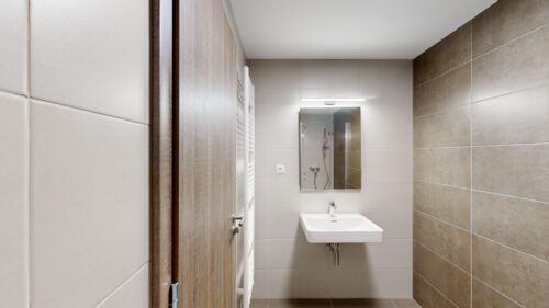 View into a modern bathroom featuring a white wall-mounted sink, rectangular mirror with a slim light, beige tiled walls, and a white towel radiator by a wooden door.