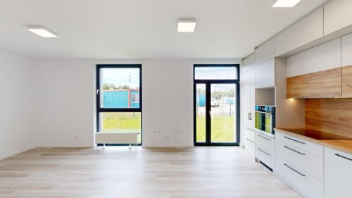Bright modern kitchen with white cabinets, wooden backsplash, and large daylight-filled windows/doors on the right side.
