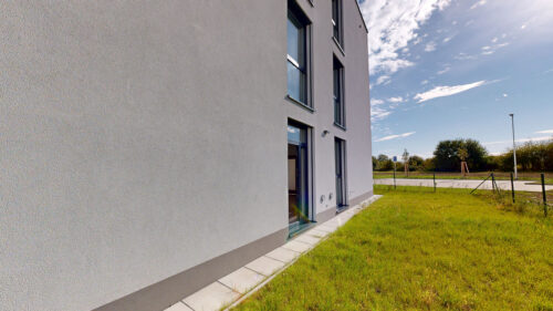 Modern building exterior with large vertical windows, beige-gray walls, and a grassy area under a blue sky.