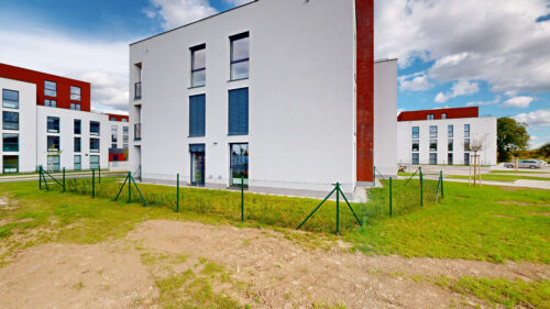 Modern white apartment buildings with red brick accents, green fencing, and a grassy foreground under a blue, partly cloudy sky