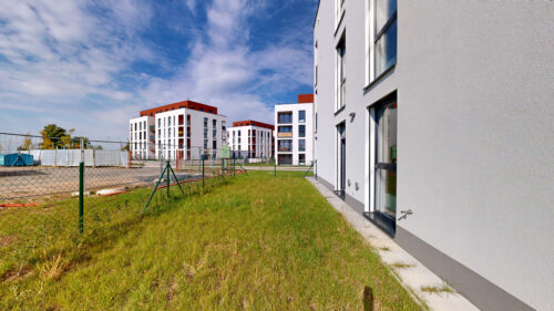 Row of white, modern apartment buildings with red accents under a blue sky, seen behind a chain-link fence and grassy lot.
