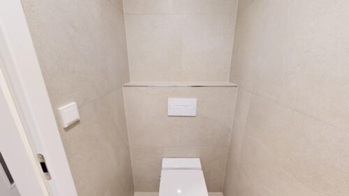 Tiny beige-tiled bathroom with a wall-hung toilet, flush plate, and a slim metal-edged shelf mid‑wall behind the toilet. A light switch is on the left wall.