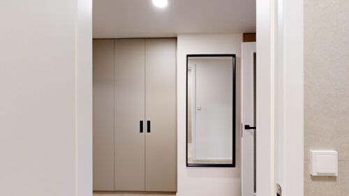 Narrow hallway with beige closet doors and a tall black-framed full‑length mirror on the right, reflecting a doorway.