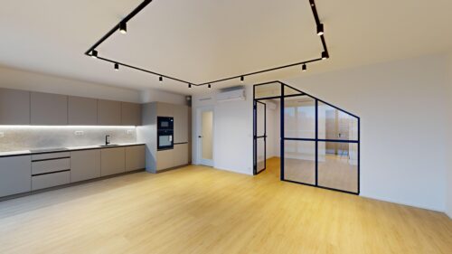 Modern open-plan kitchen with grey cabinets, built-in oven, and track lighting; glass partition with dark frame on right.