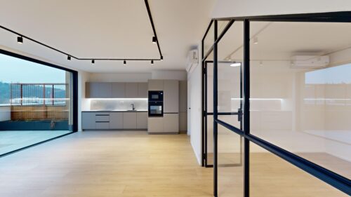 Open-plan modern kitchen with grey cabinets, built-in oven, and a large glass wall opening to an outdoor patio area outside balcony railing visible outside the glass doors.