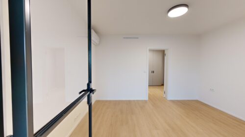 Empty modern room with light wood floor, white walls, ceiling light, and an open doorway to another room with a pair of cabinets in the distance.