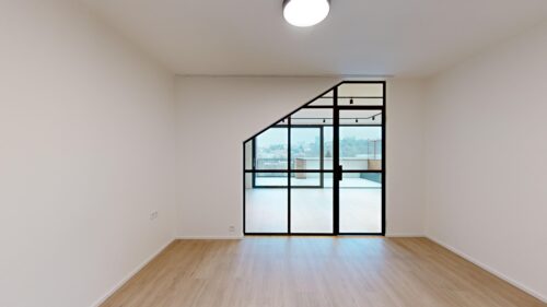 Empty modern room with light wood floors and white walls, large glass door with black frame leading to a balcony and city view outside.