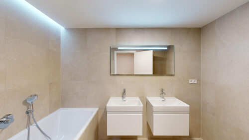 Beige-tiled bathroom with a white bathtub on the left, two square wall-mounted sinks, and a backlit rectangular mirror above them.