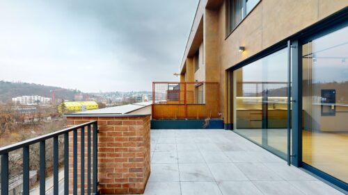Rooftop terrace of a modern building with large glass doors, brick planter, and orange metal railing; city skyline under a cloudy sky in the background.