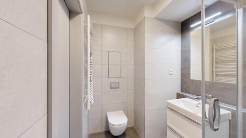 Modern bathroom with wall tiles, a wall-mounted toilet, white sink, and glass shower enclosure with a chrome handle. A white towel warmer is visible on the left.