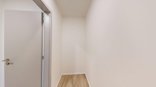 Narrow white hallway with light wood flooring and an open door on the left.