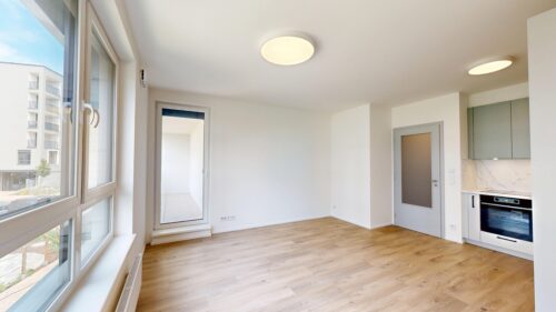Bright open-plan living area with large windows, white walls, and a light wood floor; kitchen with built-in oven on the right.
