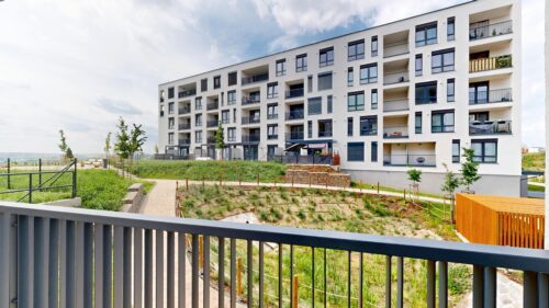 Modern white apartment building with numerous balconies, seen from a railing-lined foreground over a landscaped courtyard and walkway under a blue sky.