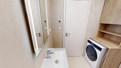 Small modern bathroom featuring a white sink with chrome faucet, beige tiles, a large white storage cabinet, and a front-loading washing machine on wooden shelves to the right.