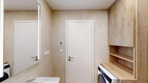 Bathroom with beige tiles, a large mirror, white doors, and a wooden shelving unit housing a washing machine at the bottom right.