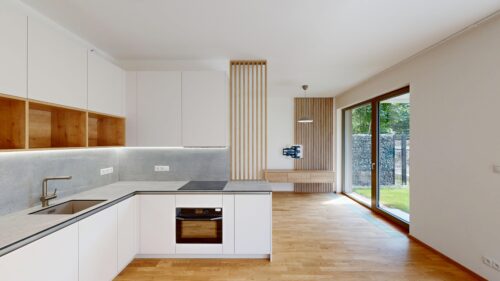 Modern white kitchen with wooden open shelves, grey countertop, and built-in oven under a light grey backsplash. On the right, a glass door opens to a green outdoor area with wooden floor. A vertical wooden slat divider separates dining area from kitchen.