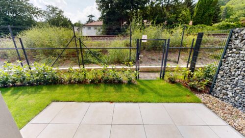 Backyard garden with a gray metal mesh fence, gate, green lawn, and planting beds along the border.