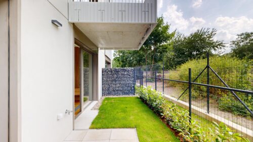 Modern house exterior with sliding glass doors, a concrete overhang, and a landscaped backyard surrounded by a metal fence.