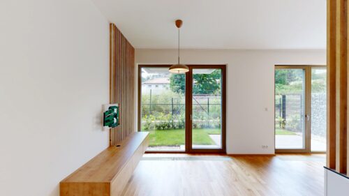 Bright modern interior with large glass doors opening to a garden, wooden bench along left wall, and a hanging pendant light.