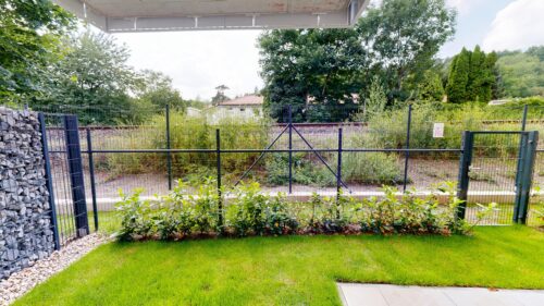 Black metal fence enclosing a garden with shrubs, trees, and a green lawn in the foreground.