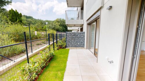 Patio of a modern apartment building with white walls, large glass doors, and a tiled floor beside a metal fence and green lawn.