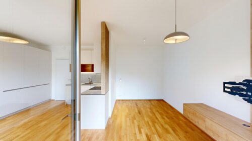 Open-plan living space with white walls, light wood floors, a glass divider, and a visible white-kitchen area through a doorway on the left; a round pendant light hangs from the ceiling.