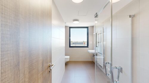 Bright modern bathroom with a glass shower enclosure, white tiles, a wall radiator, and a window at the far end; daylighted space.