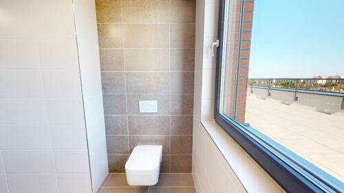 Modern bathroom with wall-hung white toilet, rectangular flush plate, and tan tiled wall next to a large window looking onto a balcony and blue sky