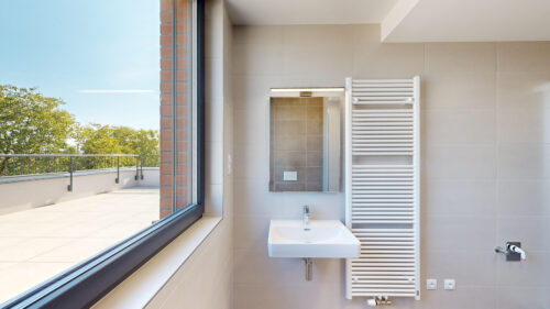 Modern bathroom interior with a wall-mounted sink, backlit mirror, and a tall white towel radiator; large window shows a balcony and trees outside.