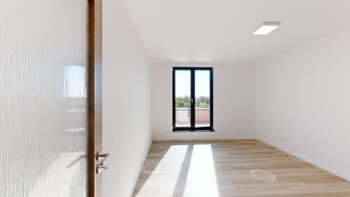 Bright empty room with light wood flooring and a large glass door/window at the far end, letting sunlight in.