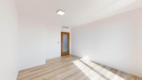 Empty white room with a wooden door, wall-mounted AC, and square ceiling light; sunlight casts shadows on light wood flooring.