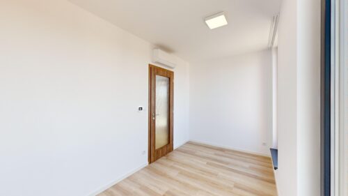 Bright, empty room with light wood flooring, a wooden frosted-glass door, and a wall-mounted air conditioner under a square ceiling light.