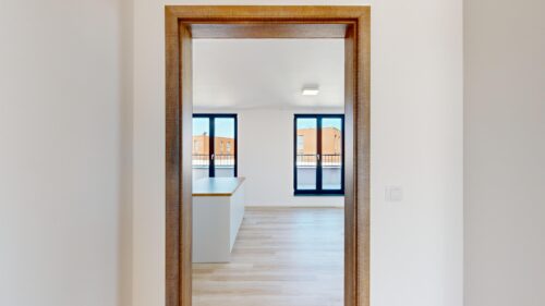 Wood-framed doorway opening into a bright room with large windows showing brick buildings outside.