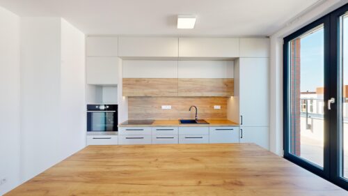 Modern minimalist kitchen with white cabinets, wood countertops, built-in oven, and a large glass door to a balcony/outdoor view.