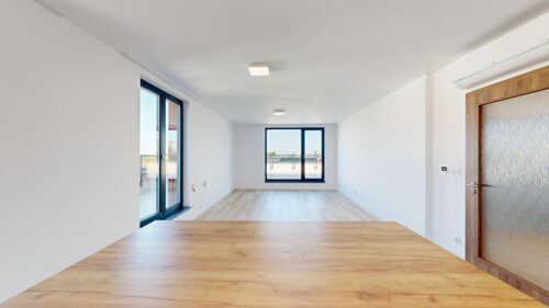 Bright empty living room with white walls, light wood floor, large sliding glass doors to a balcony, and a wooden door with frosted glass on the right.