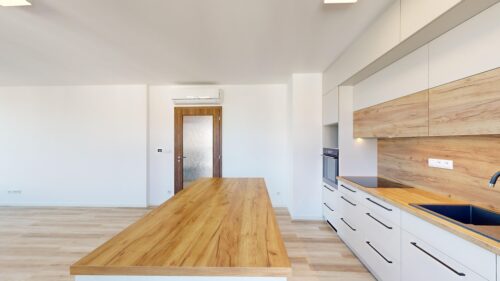 Bright modern kitchen with a long wooden island, white cabinets, and a built-in oven along a wood-paneled backsplash.