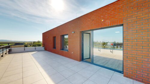 Rooftop terrace on a red brick building with large glass doors, windows, and a railing under a clear sky.