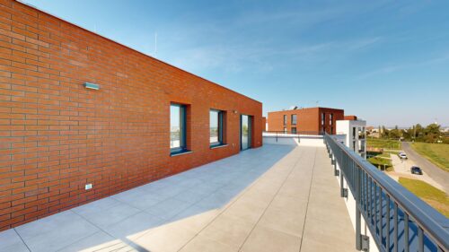 Rooftop terrace on a brick building with large windows and a metal railing, overlooking a suburban landscape under a clear blue sky.