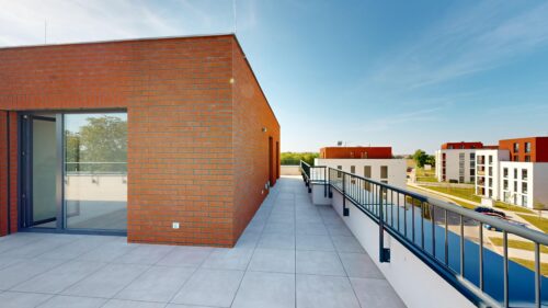 Rooftop terrace with a brick wall, glass door, and a black railing overlooking modern white and red apartment blocks on a sunny day.
