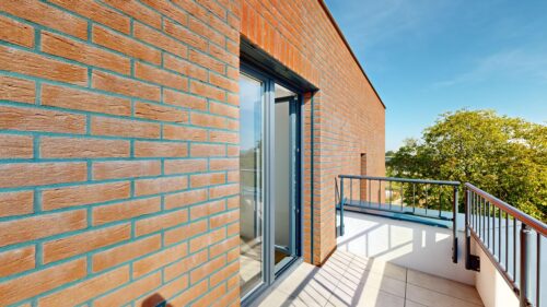 Brick exterior wall with a glass door opening to a balcony, metal railing, and a clear blue sky with trees in the background.