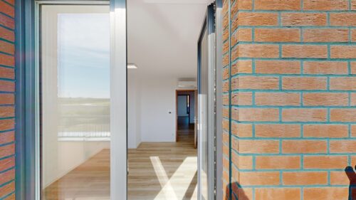 View through an open glass door to a bright interior hallway with light wood floors, brick exterior wall on the right.