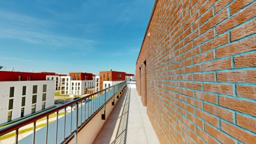 Rooftop walkway beside a tall brick wall, with a metal railing and modern white and red apartment buildings under a blue sky.
