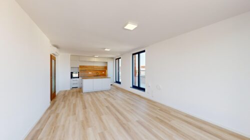 Open-plan living space with white walls, light wood floors, a centered kitchen island, and large windows letting in natural light.