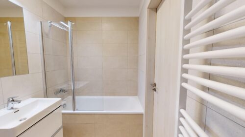 Modern bathroom with beige tiled walls, glass shower enclosure, and a white sink and tub. A white towel radiator runs along the right wall.