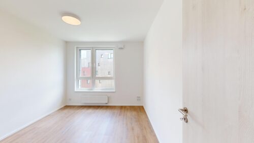 Empty bright room with white walls, light wood floor, a windowed wall at the far end, radiator below the window, and a white door ajar on the right.